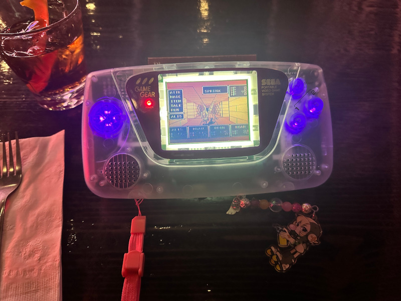 a clear sega game gear at a bar with a drink next to it, the buttons are lit up purple and it's playing Phantasy Star via the master system converter